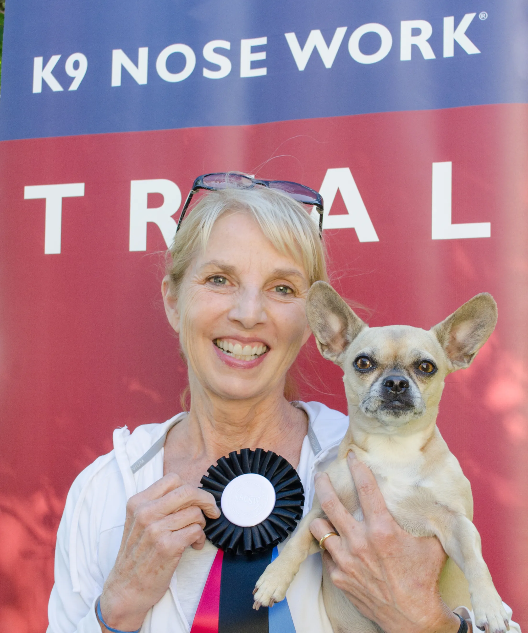 Woman holding a small dog and a ribbon at a dog event.