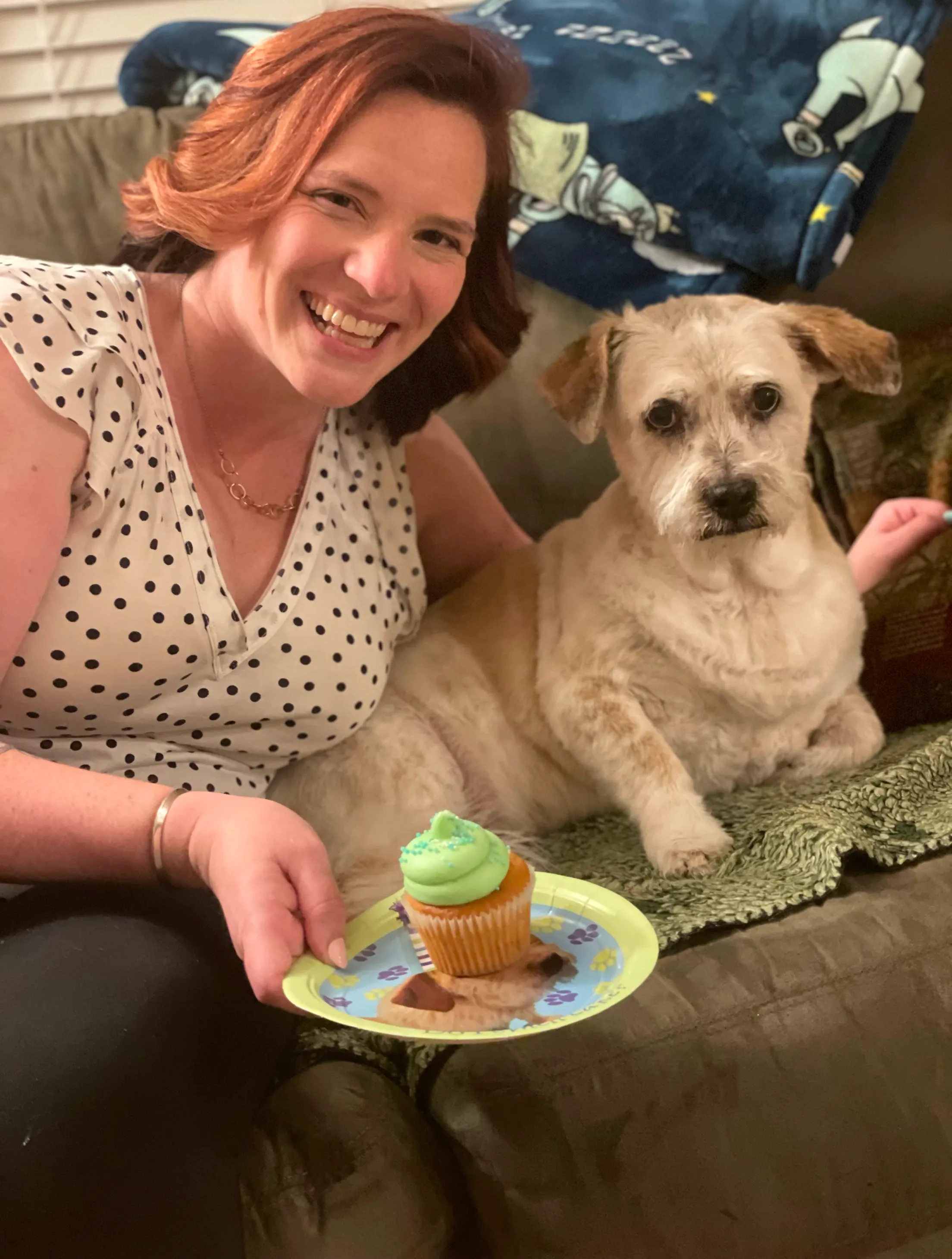 Woman smiling next to a dog sitting on a couch with a cupcake.